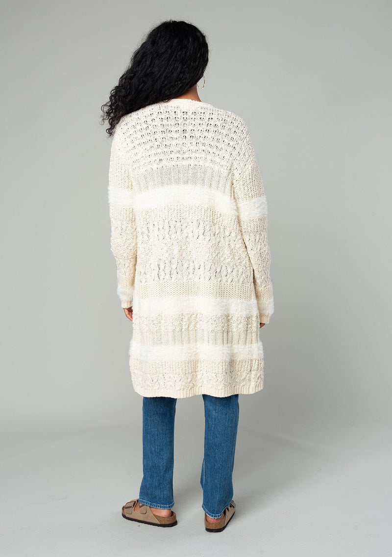 [Color: Eggshell] A back facing image of a brunette model wearing a cream colored fuzzy bohemian mid length cardigan in a textured patchwork knit. With long sleeves and an open front. 