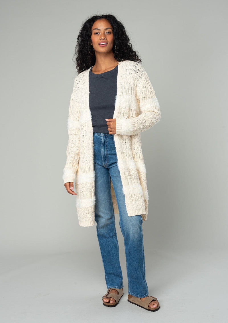 [Color: Eggshell] A front facing image of a brunette model wearing a cream colored fuzzy bohemian mid length cardigan in a textured patchwork knit. With long sleeves and an open front. 