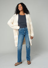 [Color: Eggshell] A full body front facing image of a brunette model wearing a cream colored fuzzy bohemian mid length cardigan in a textured patchwork knit. With long sleeves and an open front. 