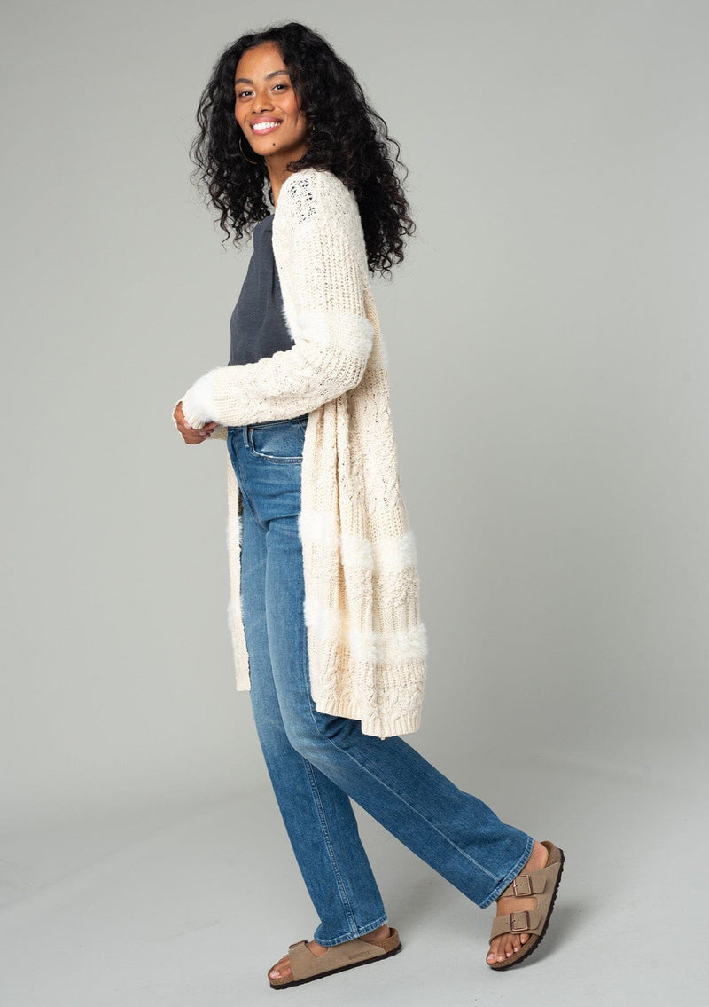 [Color: Eggshell] A side facing image of a brunette model wearing a cream colored fuzzy bohemian mid length cardigan in a textured patchwork knit. With long sleeves and an open front. 