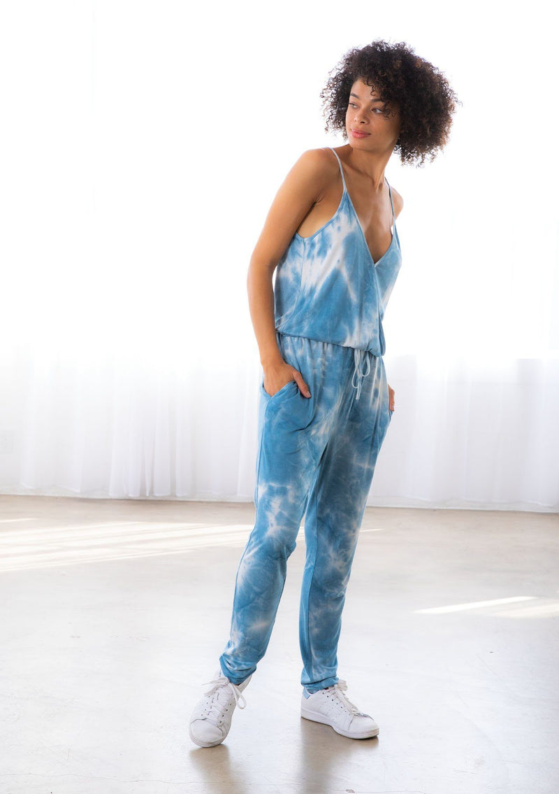 [Color: Blue] A model wearing a sleeveless blue tie dye knit jumpsuit. With a long tapered leg, a surplice v neckline, side pockets, and a drawstring waist.