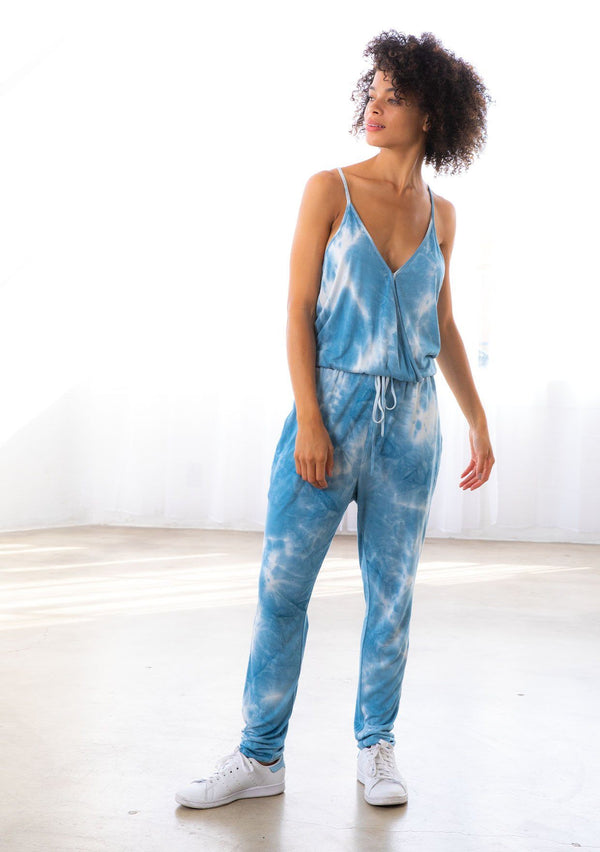 [Color: Blue] A model wearing a sleeveless blue tie dye knit jumpsuit. With a long tapered leg, a surplice v neckline, side pockets, and a drawstring waist.