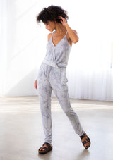 [Color: Grey] A model wearing a sleeveless grey tie dye knit jumpsuit. With a long tapered leg, a surplice v neckline, side pockets, and a drawstring waist.