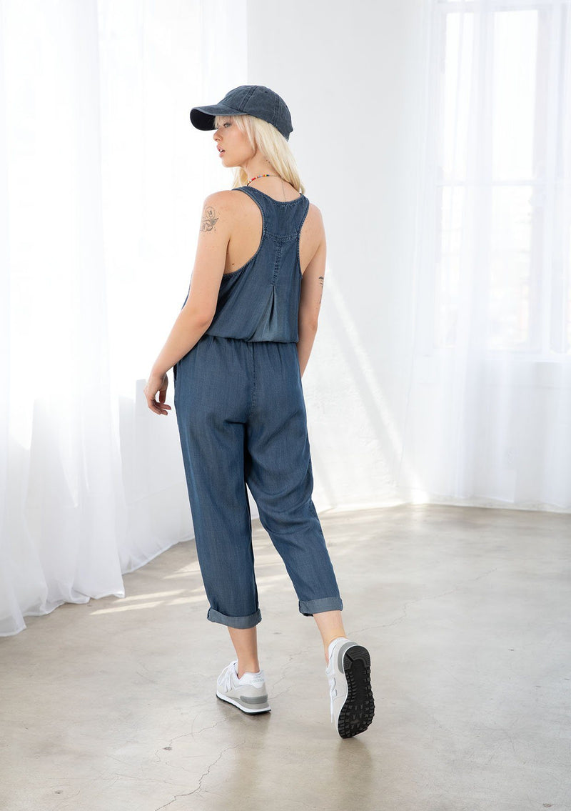 [Color: Midnight Wash] A blond model wearing a denim blue classic jumpsuit made from eco friendly Tencel. Featuring a wide leg with a cuffed hem, essential side pockets, and a button front.
