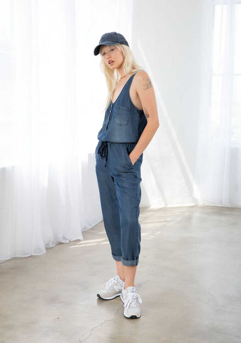[Color: Midnight Wash] A blond model wearing a denim blue classic jumpsuit made from eco friendly Tencel. Featuring a wide leg with a cuffed hem, essential side pockets, and a button front.