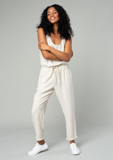 [Color: Natural] A full body front facing image of a brunette model wearing a classic bohemian one piece natural jumpsuit. With a racer back, a button front top, side pockets, a cuffed wide leg, a scoop neckline, and an elastic waist with drawstring. 