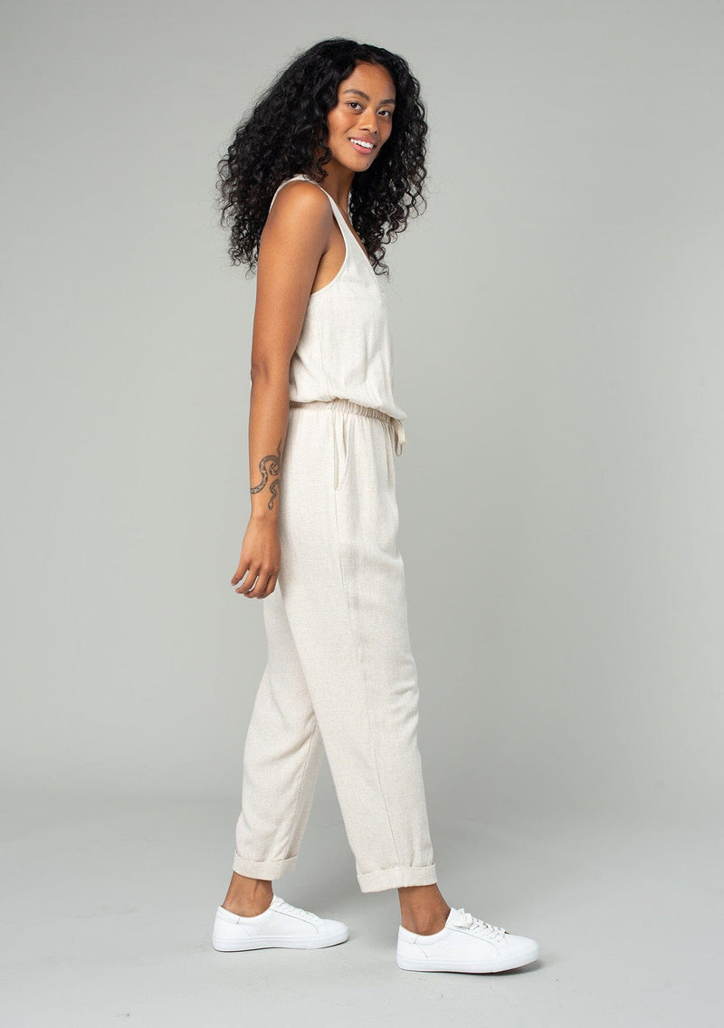 [Color: Natural] A side facing image of a brunette model wearing a classic bohemian one piece natural jumpsuit. With a racer back, a button front top, side pockets, a cuffed wide leg, a scoop neckline, and an elastic waist with drawstring. 