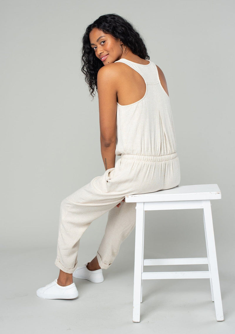 [Color: Natural] A sitting back facing image of a brunette model wearing a classic bohemian one piece natural jumpsuit. With a racer back, a button front top, side pockets, a cuffed wide leg, a scoop neckline, and an elastic waist with drawstring. 