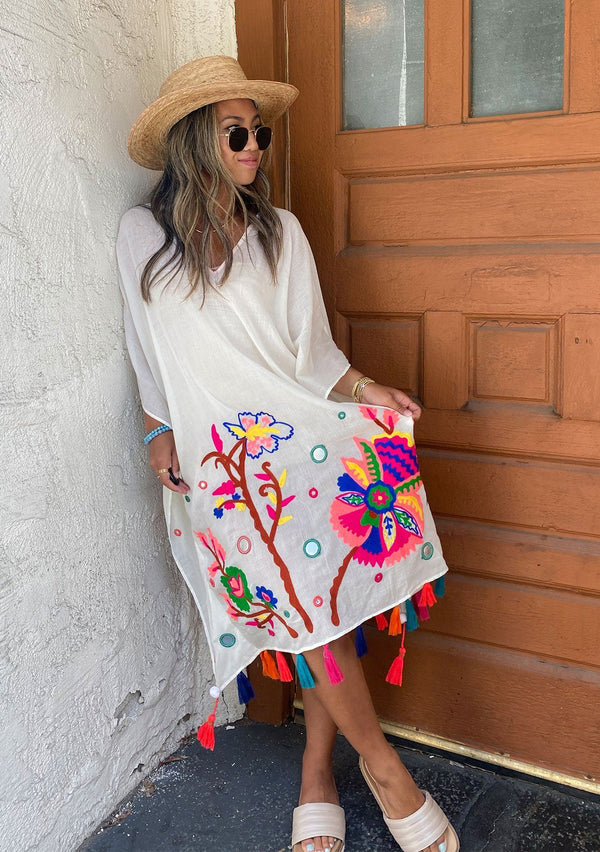 [Color: Off White] A brunette woman standing outside wearing a mid length sheer caftan with bold embroidered detail and colorful tassel trim.