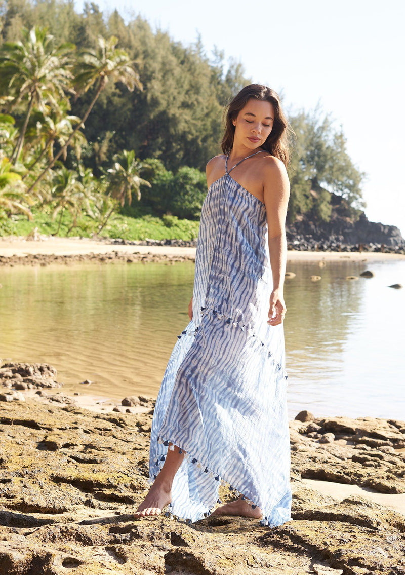 [Color: Blue] A model wearing a sheer blue tie dye halter maxi dress. Featuring a strappy open back, a tassel trim throughout, and a front keyhole detail. 