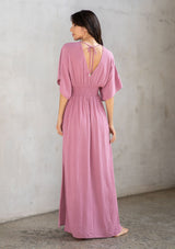 [Color: Smokey Orchid] A model wearing a resort ready light purple maxi dress. With half length kimono sleeves, a plunging v neckline, a smocked elastic empire waist, side slits, and an open back with tie closure.