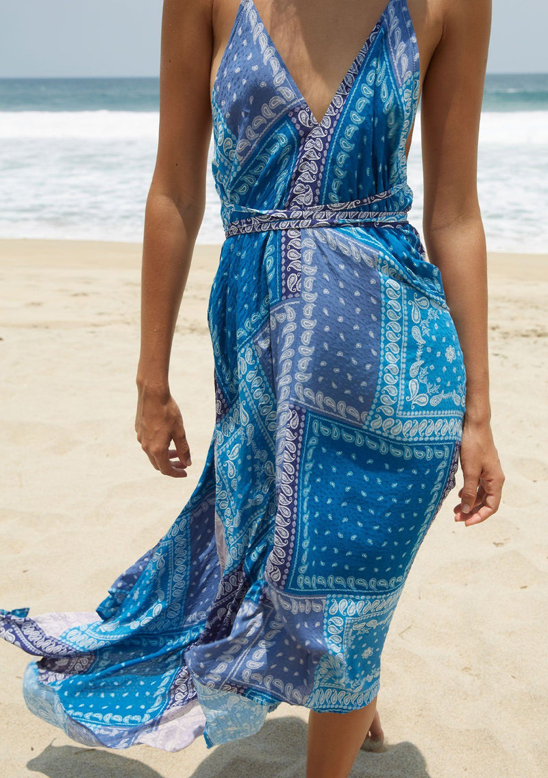 [Color: Purple/Blue] A model wearing a halter maxi dress in a purple and blue paisley bandana patchwork print. With a deep v neckline, an open back, and long straps that can be tied in multiple ways.