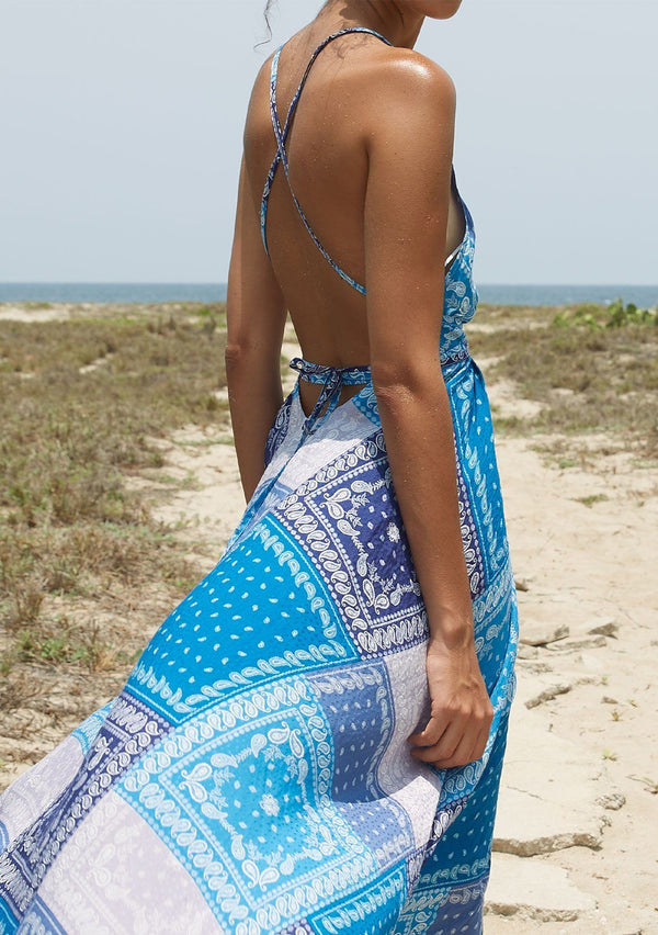 [Color: Purple/Blue] A model wearing a halter maxi dress in a purple and blue paisley bandana patchwork print. With a deep v neckline, an open back, and long straps that can be tied in multiple ways.