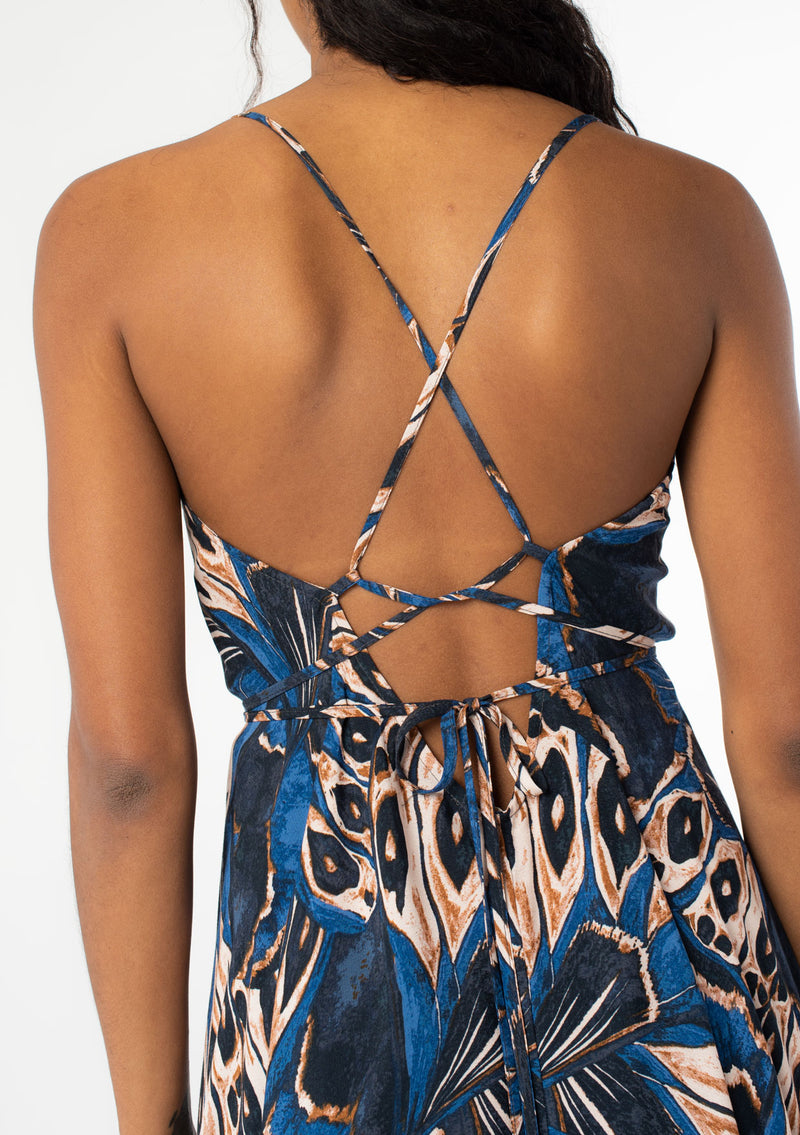 [Color: Cobalt/Tan] A close up back facing image of a black model wearing a blue and tan butterfly wing print mid length halter dress with long straps that can be tied multiple ways. 