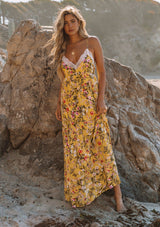 [Color: Corn Combo] A woman standing on the beach wearing a yellow floral maxi slip dress. With a tiered skirt, a lace trim v neckline and racer back, and adjustable spaghetti straps.