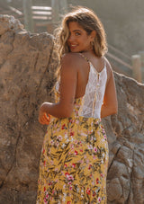 [Color: Corn Combo] A woman standing on the beach wearing a yellow floral maxi slip dress. With a tiered skirt, a lace trim v neckline and racer back, and adjustable spaghetti straps.