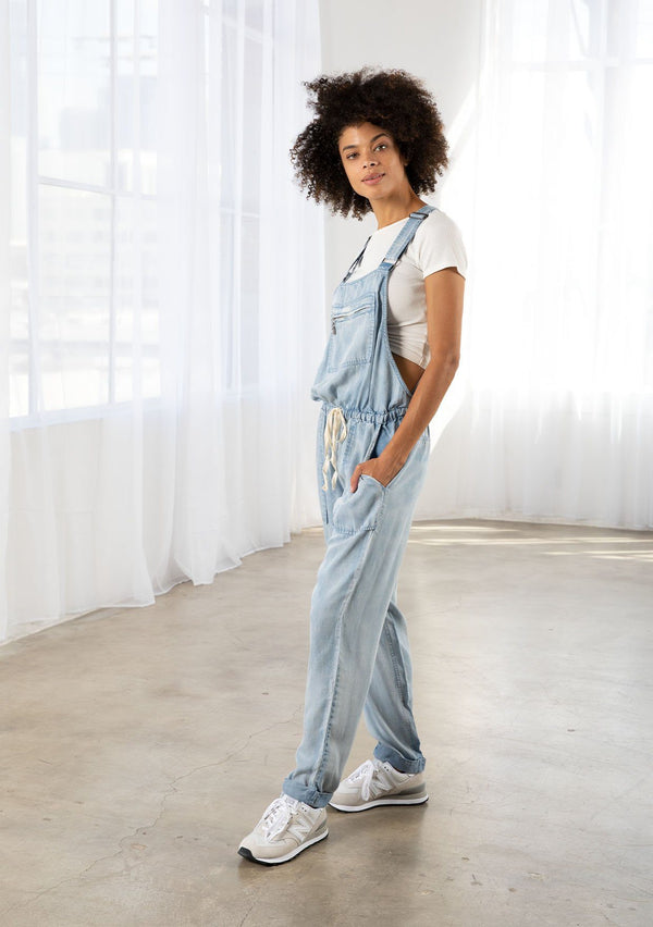 [Color: Reagan Wash] A model wearing classic light wash overalls. With a drawstring waist, side pockets, adjustable straps, a full length leg, and a zippered front pocket.