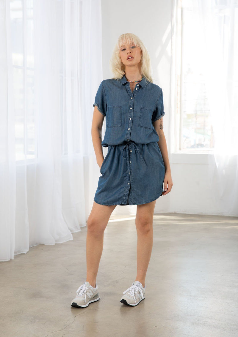 [Color: Vintage Wash] A blond model wearing a casual denim blue short sleeve shirt dress. Featuring an adjustable drawstring waist for definition, side pockets, and a chic shirttail hemline.