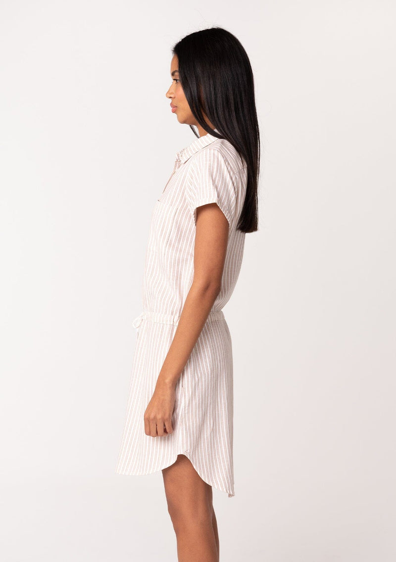 [Color: White/Sand] A side facing image of a brunette model wearing a classic mini shirtdress in a white and sand stripe. With short cuffed sleeves, side pockets, a shirttail hemline, a button front, and a drawstring waist.