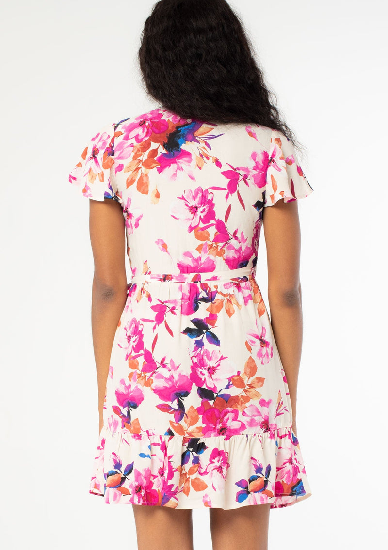 [Color: Ivory/Fuchsia] A back facing image of a black model with long dark wavy hair wearing a white and pink watercolor floral print mini wrap dress. With short flutter sleeves, a side tie closure, and a ruffled hemline. 