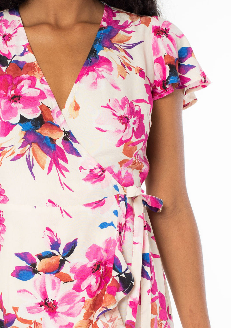 [Color: Ivory/Fuchsia] A close up front facing image of a black model with long dark wavy hair wearing a white and pink watercolor floral print mini wrap dress. With short flutter sleeves, a side tie closure, and a ruffled hemline. 