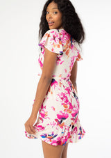 [Color: Ivory/Fuchsia] A close up side facing image of a black model with long dark wavy hair wearing a white and pink watercolor floral print mini wrap dress. With short flutter sleeves, a side tie closure, and a ruffled hemline. 
