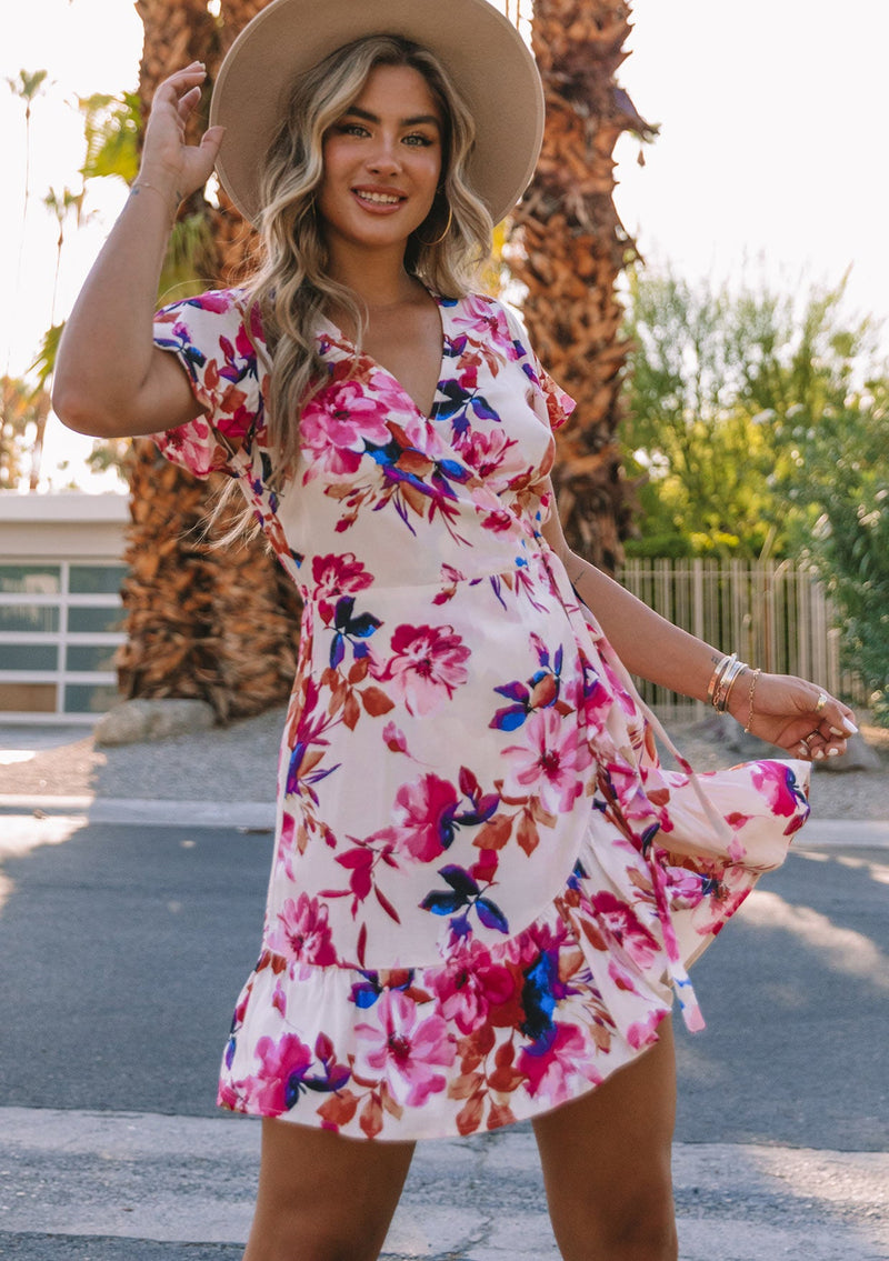 [Color: Ivory/Fuchsia] A front facing image of a model with long blonde wavy hair wearing a white and pink watercolor floral print mini wrap dress. With short flutter sleeves, a side tie closure, and a ruffled hemline.
