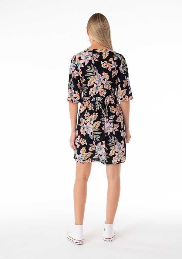 [Color: Black/Pink] A back facing image of a blonde model wearing a lightweight spring mini dress in a pink tropical floral print. With half length short sleeves, a deep v neckline, an empire waist, and a flowy, relaxed fit. 
