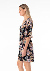 [Color: Black/Pink] A side facing image of a blonde model wearing a lightweight spring mini dress in a pink tropical floral print. With half length short sleeves, a deep v neckline, an empire waist, and a flowy, relaxed fit. 