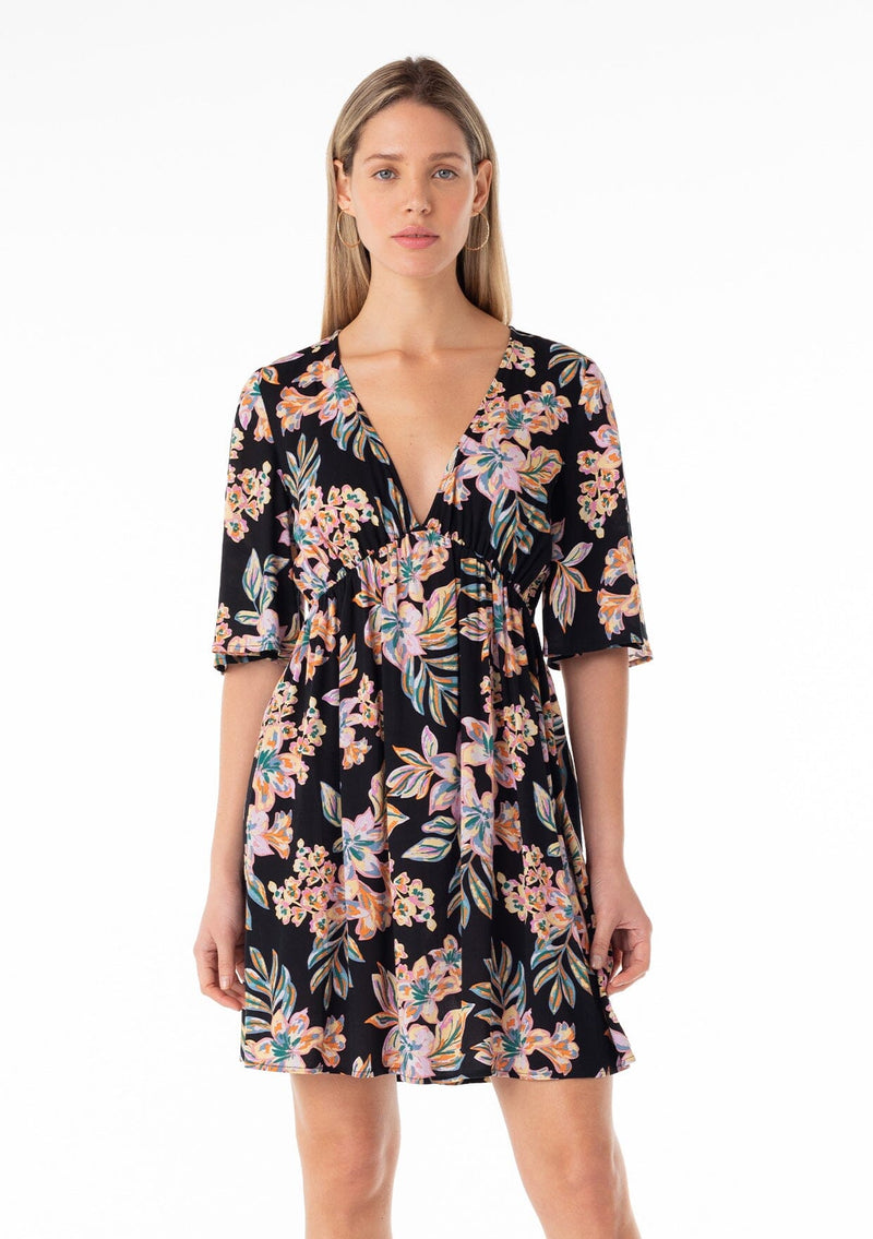 [Color: Black/Pink] A front facing image of a blonde model wearing a lightweight spring mini dress in a pink tropical floral print. With half length short sleeves, a deep v neckline, an empire waist, and a flowy, relaxed fit. 