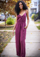 [Color: Burgundy] A woman wearing a one piece jumpsuit. Featuring a wide leg, a front keyhole detail, an elastic cinched waist, and side pockets.