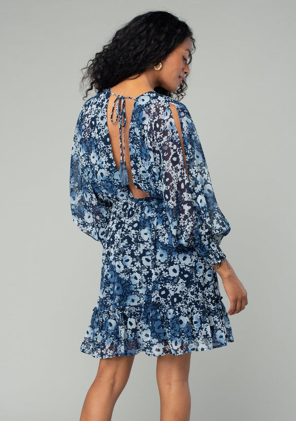 [Color: Indigo/Blue] A back facing image of a brunette model wearing a best selling bohemian chiffon mini dress in a blue floral print. With long split sleeves, a ruffle trimmed tiered skirt, a smocked elastic waist, an open back with tassel tie closure, and a v neckline. 