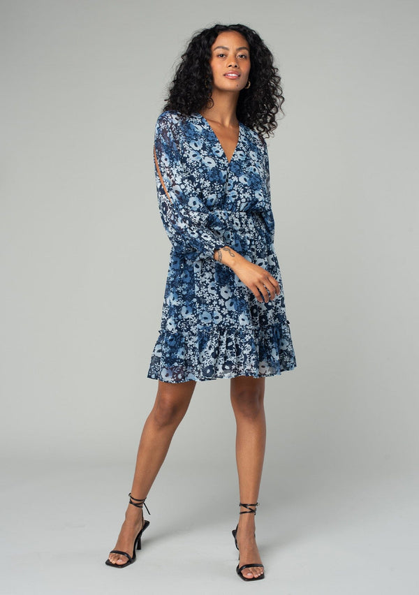 [Color: Indigo/Blue] A front facing image of a brunette model wearing a best selling bohemian chiffon mini dress in a blue floral print. With long split sleeves, a ruffle trimmed tiered skirt, a smocked elastic waist, an open back with tassel tie closure, and a v neckline. 
