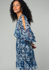 [Color: Indigo/Blue] A side facing image of a brunette model wearing a best selling bohemian chiffon mini dress in a blue floral print. With long split sleeves, a ruffle trimmed tiered skirt, a smocked elastic waist, an open back with tassel tie closure, and a v neckline. 