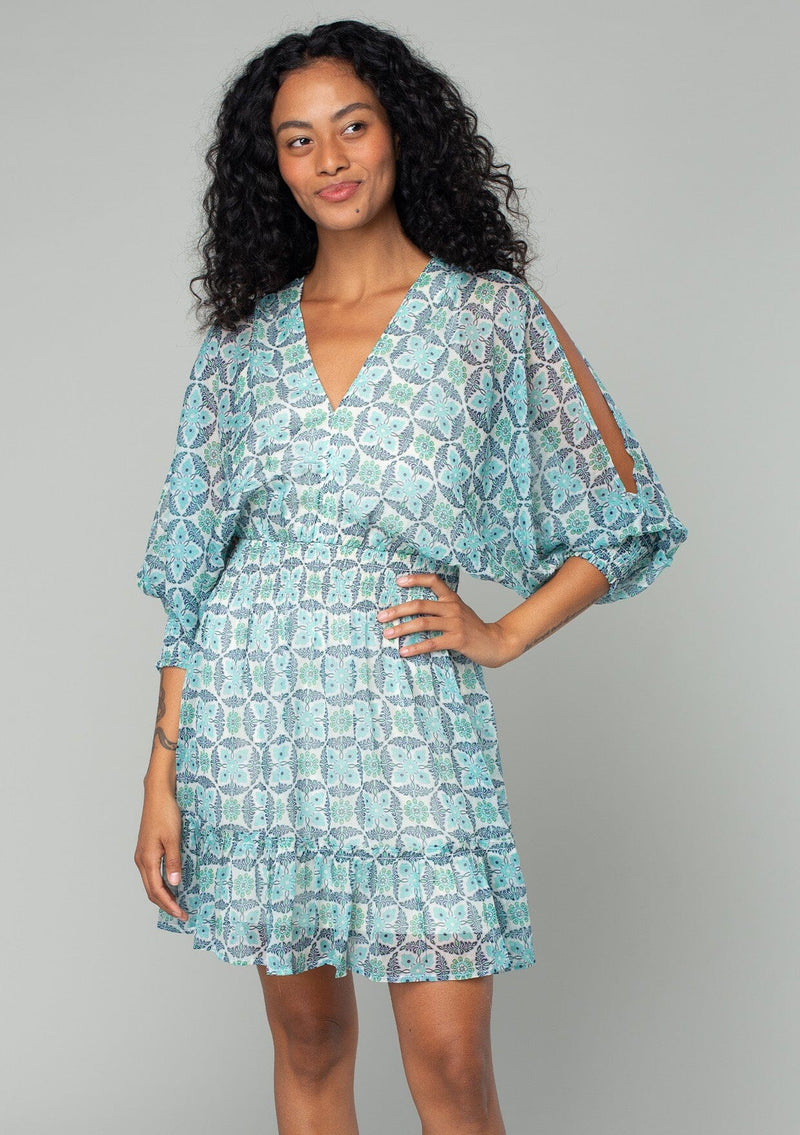 [Color: Cream/Navy] A front facing image of a brunette model wearing a best selling chiffon bohemian mini dress in a cream and navy blue geometric floral print. With long voluminous split sleeves, a smocked elastic waist, a ruffle trimmed tiered skirt, a v neckline, and an open back with tassel tie closure. 