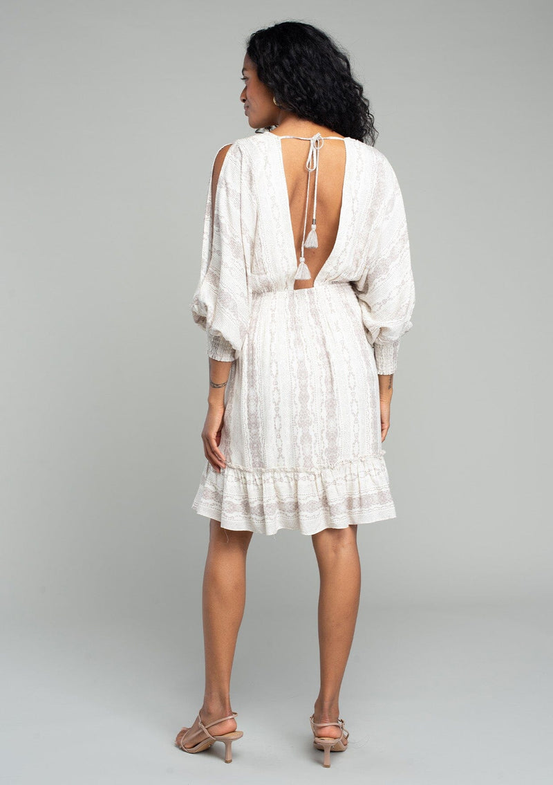 [Color: Ivory/Grey] A back facing image of a brunette model wearing a best selling off white bohemian mini dress with a grey bohemian mixed print. With long voluminous split sleeves, a v neckline, a smocked elastic waist, a ruffle trimmed tiered skirt, and an open back with tassel tie closure.