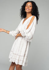 [Color: Ivory/Grey] A side facing image of a brunette model wearing a best selling off white bohemian mini dress with a grey bohemian mixed print. With long voluminous split sleeves, a v neckline, a smocked elastic waist, a ruffle trimmed tiered skirt, and an open back with tassel tie closure.