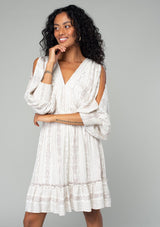 [Color: Ivory/Grey] A half body front facing image of a brunette model wearing a best selling off white bohemian mini dress with a grey bohemian mixed print. With long voluminous split sleeves, a v neckline, a smocked elastic waist, a ruffle trimmed tiered skirt, and an open back with tassel tie closure.