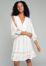 [Color: Ivory/Grey] A front facing image of a brunette model wearing a best selling off white bohemian mini dress with a grey bohemian mixed print. With long voluminous split sleeves, a v neckline, a smocked elastic waist, a ruffle trimmed tiered skirt, and an open back with tassel tie closure.