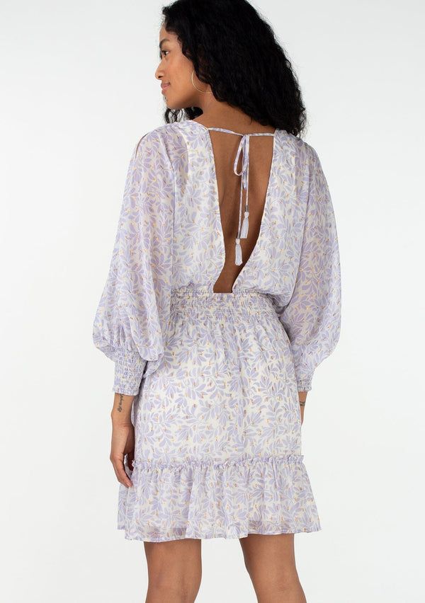 [Color: Natural/Lilac] A back facing image of a brunette model wearing a best selling mini dress in a metallic clip dot chiffon, designed in a purple leaf print. With long split sleeves, a flowy ruffle trimmed tiered skirt, a v neckline, an open back with tassel tie closure, and a smocked elastic waist. 