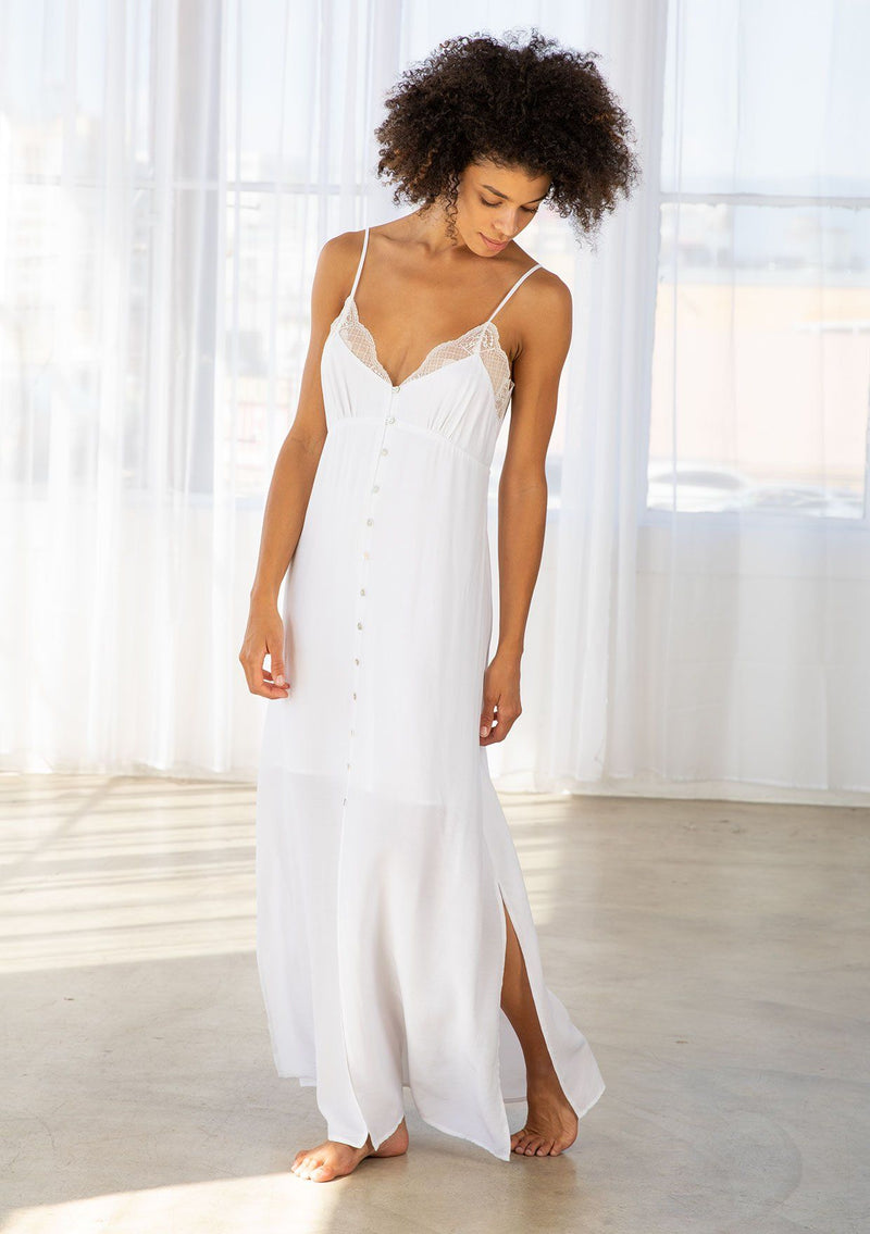 [Color: Chalk] A model wearing a soft and silky white maxi slip dress. With a lace trimmed v neckline, a button front, an empire waist, adjustable spaghetti straps, and a tie back waist detail. 