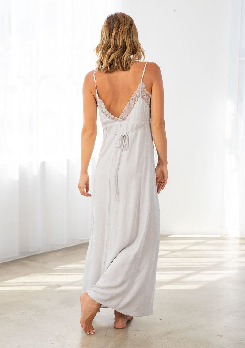 [Color: Silver] A model wearing a silver maxi slip dress in textured clip dot. With a lace trim v neckline in front and back, a self covered button front, a front slit, and a waist defining tie in the back. 