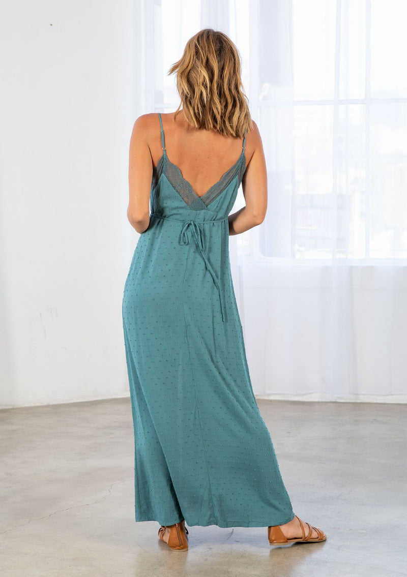 [Color: Dusty Teal] A model wearing a teal maxi slip dress in textured clip dot. With a lace trim v neckline in front and back, a self covered button front, a front slit, and a waist defining tie in the back. 