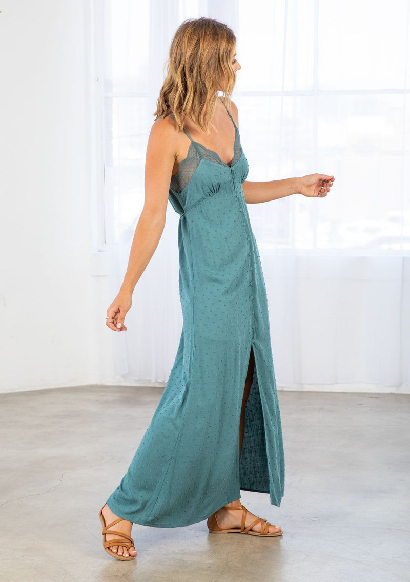 [Color: Dusty Teal] A model wearing a teal maxi slip dress in textured clip dot. With a lace trim v neckline in front and back, a self covered button front, a front slit, and a waist defining tie in the back. 
