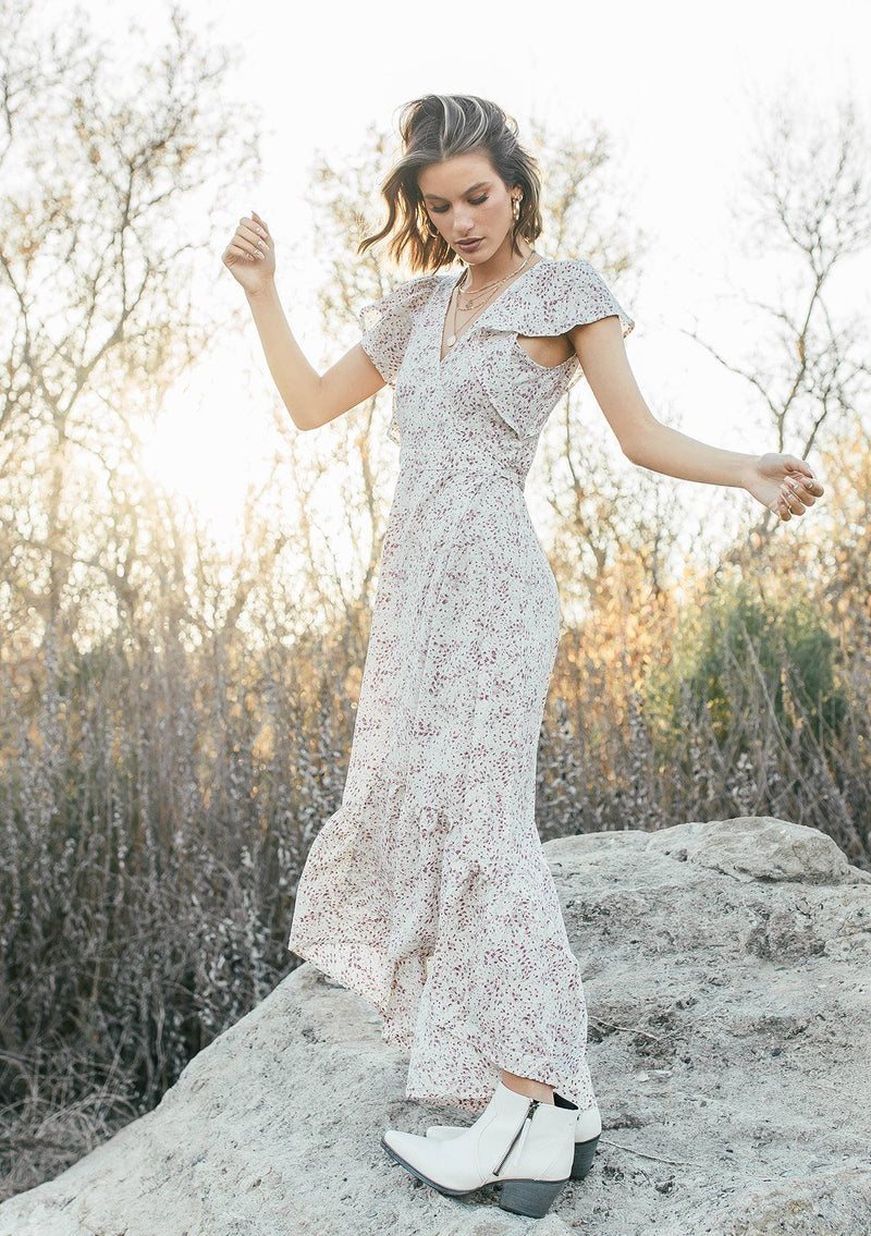 [Color: Ivory/Orchid] A woman standing on a rock in a field wearing an ultra feminine maxi wrap dress in a small floral print. Featuring a flirty short flutter sleeve and tiered skirt with a ruffled hem.