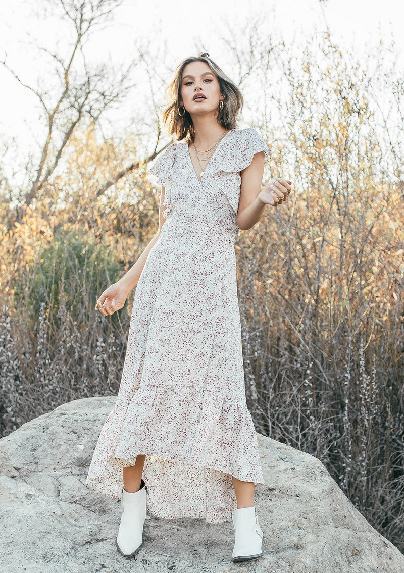[Color: Ivory/Orchid] A woman standing on a rock in a field wearing an ultra feminine maxi wrap dress in a small floral print. Featuring a flirty short flutter sleeve and tiered skirt with a ruffled hem.