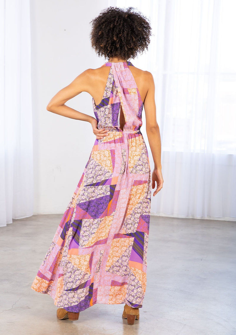 [Color: Pink/Coral] A model wearing a halter maxi dress in a geometric floral patchwork print. With an elastic waist, side pockets, a v neckline, and a cross back detail. 