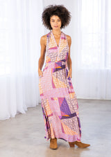 [Color: Pink/Coral] A model wearing a halter maxi dress in a geometric floral patchwork print. With an elastic waist, side pockets, a v neckline, and a cross back detail. 