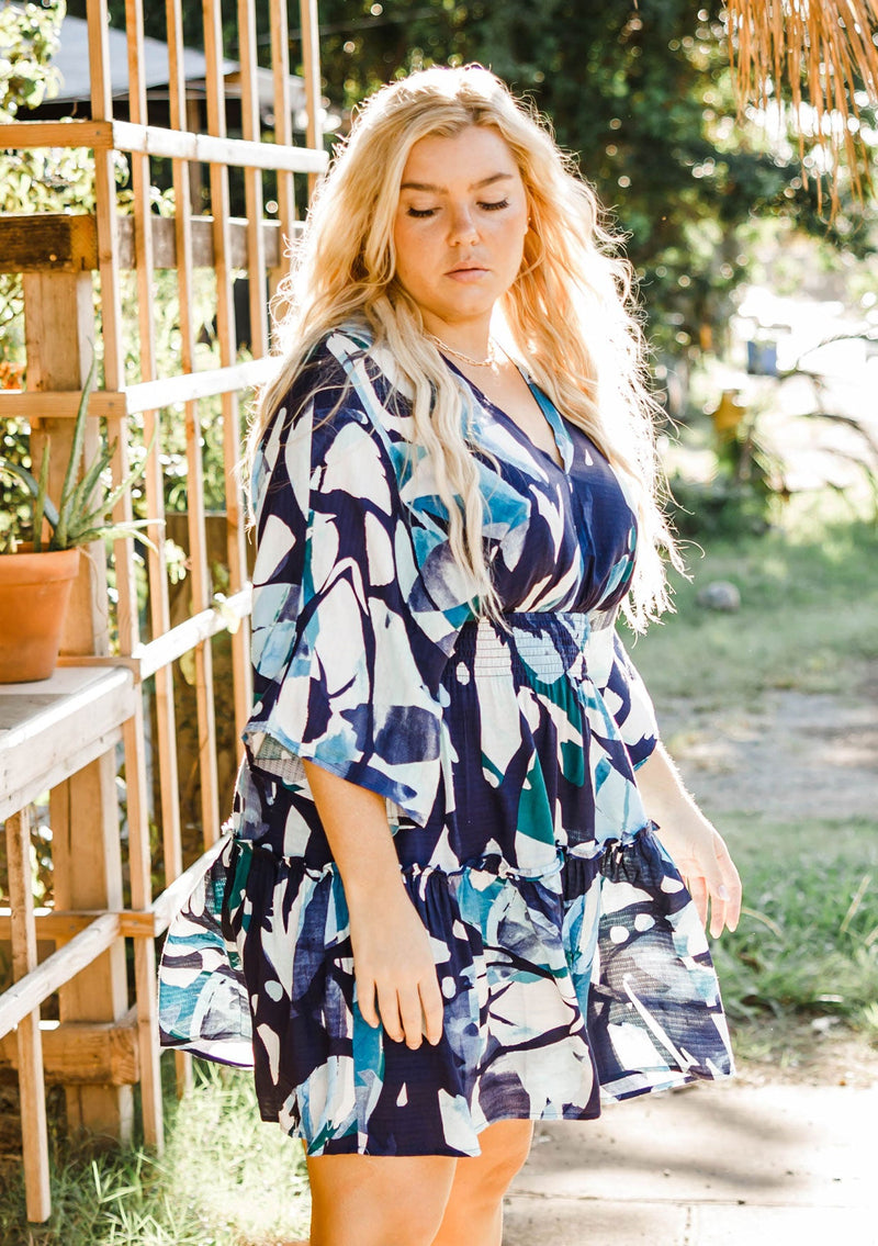 [Color: Navy/Teal] A model wearing a navy and teal bohemian watercolor print mini dress. With half length kimono sleeves, a smocked elastic waist, tiered skirt, and open tie back detail.