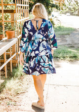 [Color: Navy/Teal] A model wearing a navy and teal bohemian watercolor print mini dress. With half length kimono sleeves, a smocked elastic waist, tiered skirt, and open tie back detail.
